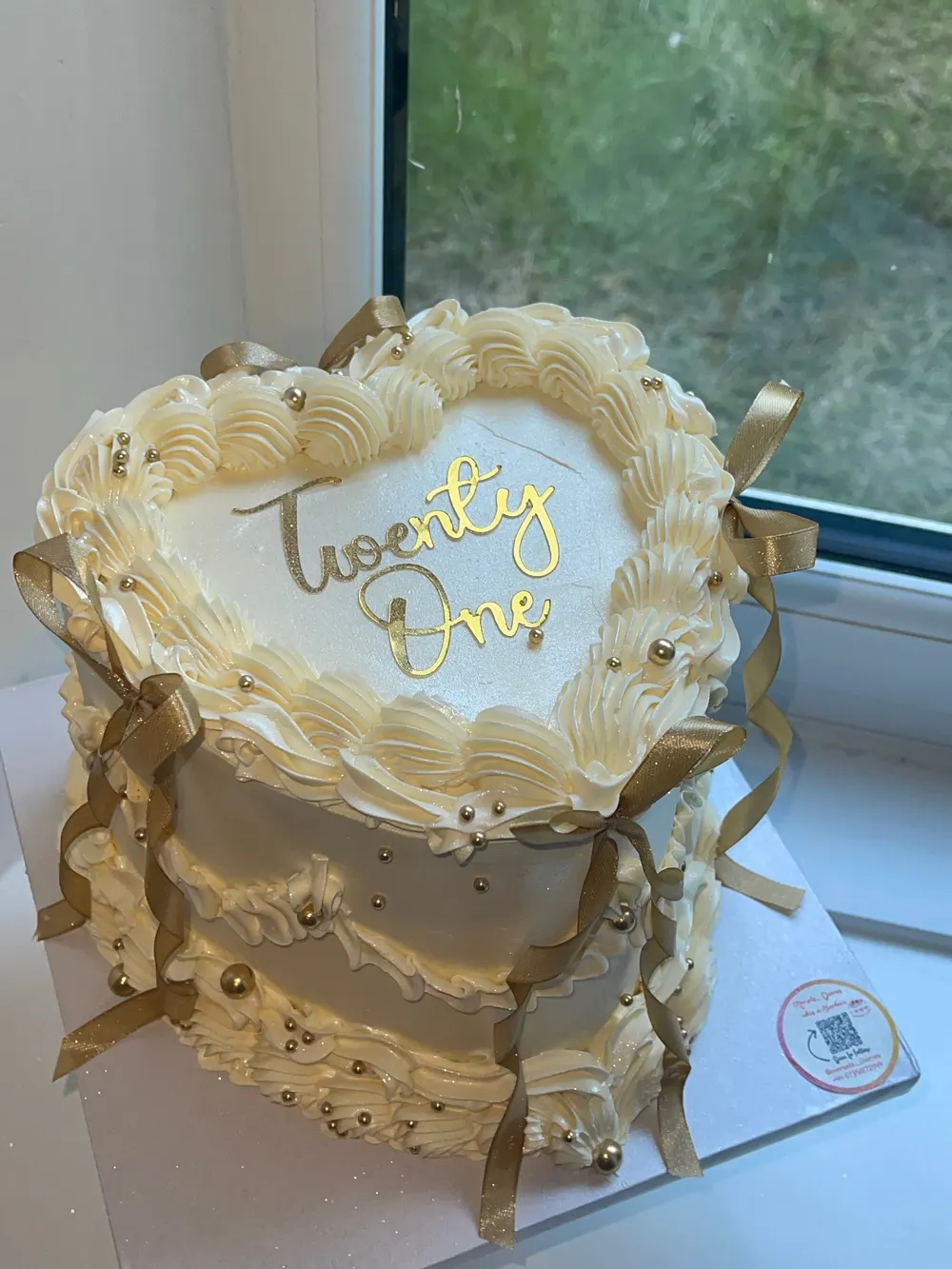 A cream heart cake with vintage piping, gold bows and a gold "Twenty One" topper on a sunny windowsill
